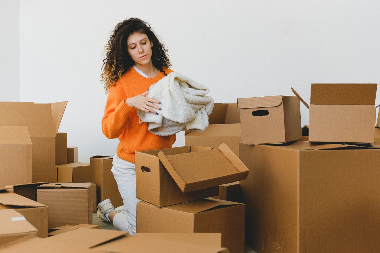 Services Concentrated young lady with curly hair standing on knees on floor and packing clothes into carton boxes before moving into new flat
